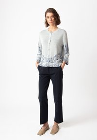 Light gray cardigan with blue floral pattern at the bottom, round neckline, and buttons. Paired with black trousers and beige flat shoes.