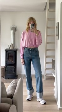 Pink cropped sweatshirt with long sleeves, high-waisted light blue jeans featuring frayed hems, and white sneakers. Simple home interior.