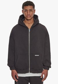 Sort zip-hoodie med en stor hætte, frontlommer, ribbede manchetter og et tekstureret stof. Har et lille logo på den nederste venstre side.