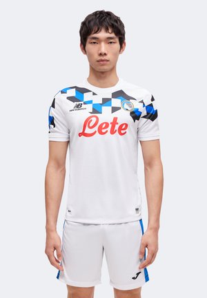 Homme portant un maillot de football blanc avec des motifs géométriques bleus et noirs et un short blanc, debout devant un fond uni.