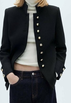 Zwarte op maat gemaakte blazer met een hoge hals, voorzien van gouden knopen aan de voorkant en op de manchetten, gecombineerd met een witte geribbelde coltrui en donkere jeans.