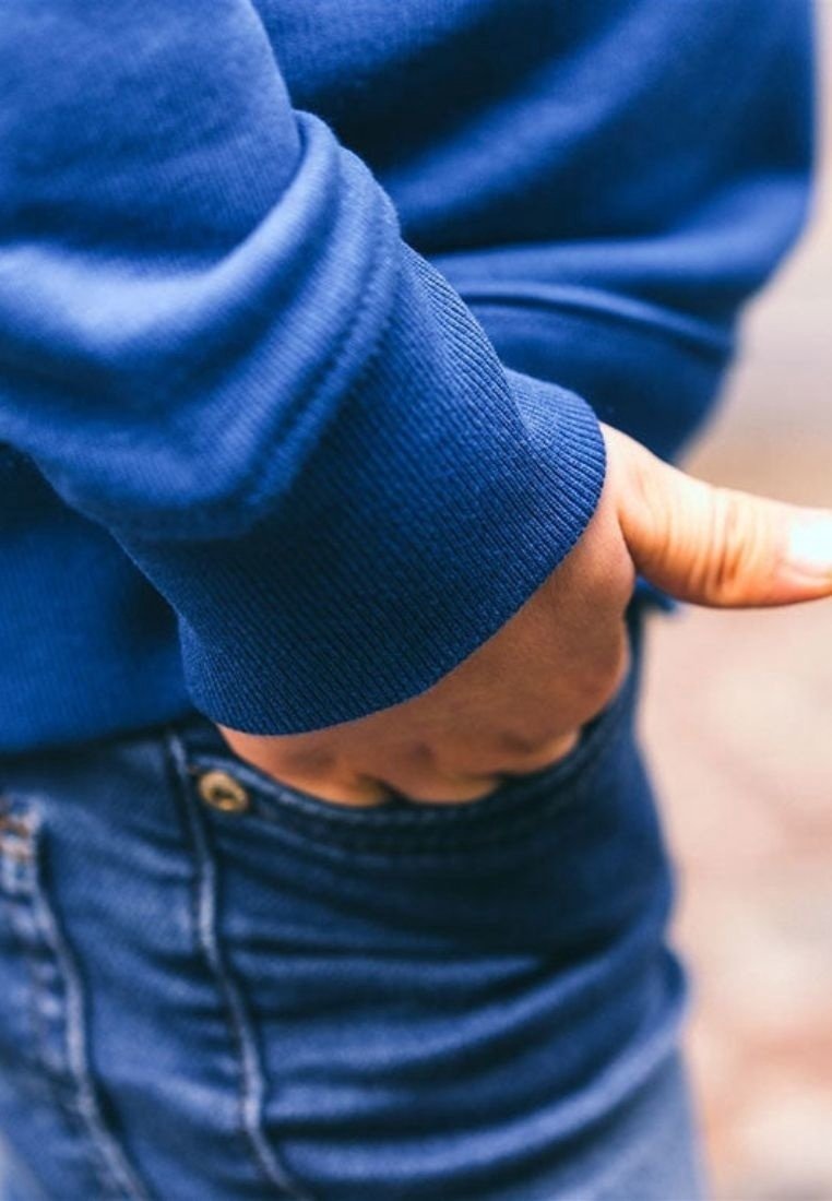 Pull bleu foncé avec poignets côtelés, comportant une main glissée dans une poche de jean en denim bleu clair. Texture douce et coupe décontractée.