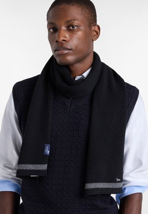 Sciarpa nera lavorata a maglia con una texture liscia, caratterizzata da accenti a righe grigie e un piccolo etichetta con logo. Indossata sopra un maglione blu navy e una camicia bianca.