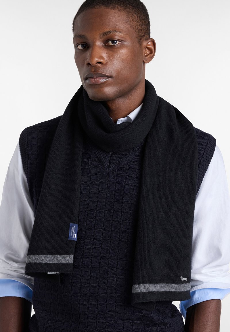 Sciarpa nera lavorata a maglia con una texture liscia, caratterizzata da accenti a righe grigie e un piccolo etichetta con logo. Indossata sopra un maglione blu navy e una camicia bianca.