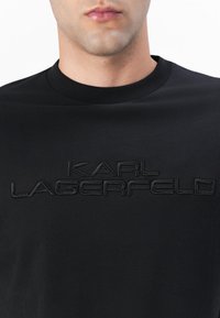 Fekete pamut póló, amelynek kerek nyaka van, és a mellkasán ellentétes textúrával feltűntetett "KARL LAGERFELD" felirattal rendelkezik.