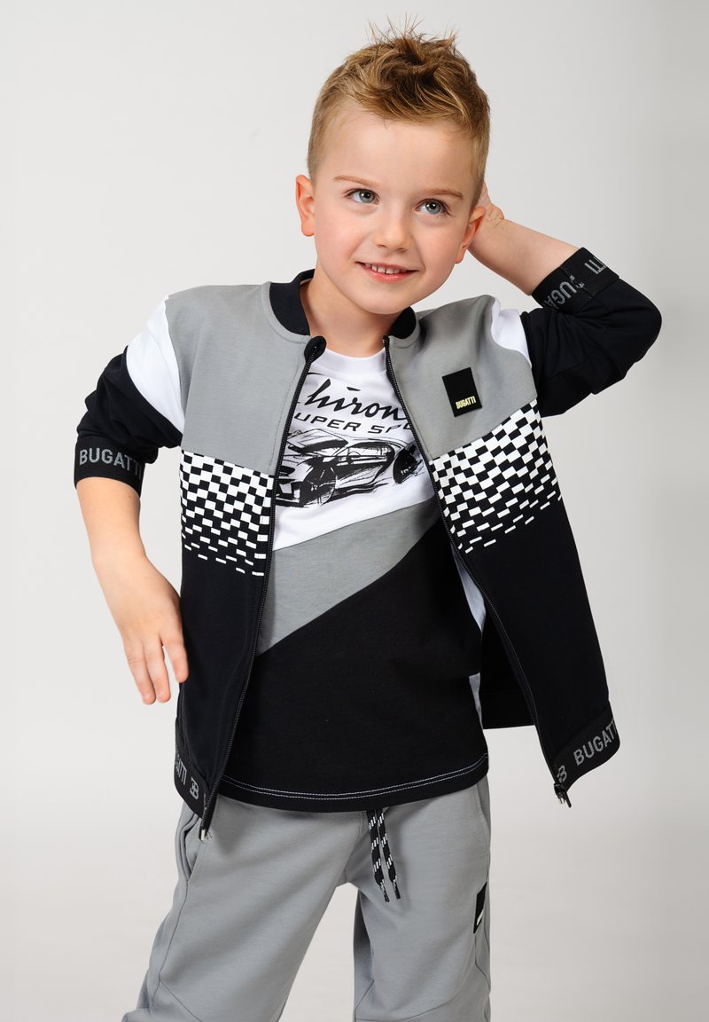 Completo sportivo da ragazzo con giacca zip color block grigia, nera e bianca, caratterizzata da un motivo a scacchi, abbinata a pantaloni grigi.