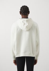 Hoodie u boji krem, izrađen od mekanog materijala. Ima opušten kroj, rebraste manžetne i šav straga.