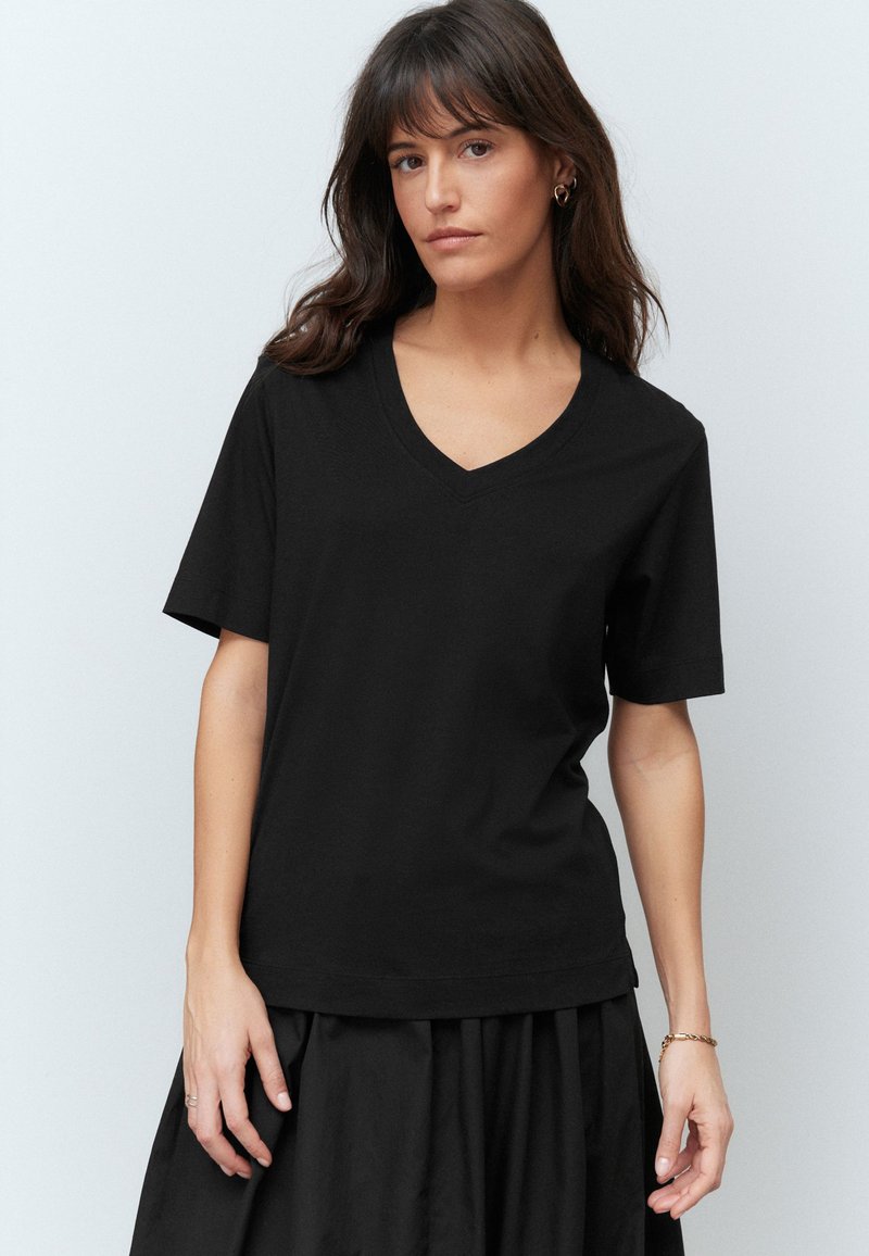 Femme aux cheveux foncés portant un t-shirt noir à manches courtes et col en V ainsi qu'une jupe noire, debout devant un fond clair uni.