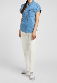 Light blue denim short-sleeve shirt with two chest pockets, button-up front, paired with white slim-fit pants and white sneakers.