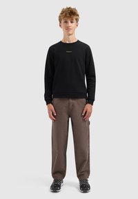 Tiener die rechtop staat en een zwarte sweatshirt, bruine wijde broek en zwarte sportschoenen draagt, tegen een effen achtergrond.