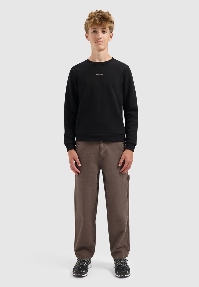 Tiener die rechtop staat en een zwarte sweatshirt, bruine wijde broek en zwarte sportschoenen draagt, tegen een effen achtergrond.