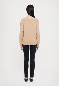 Suéter de punto beige con dobladillo y puños acanalados, combinado con jeans negros ajustados y zapatos negros sin cordones, visto desde la parte posterior.