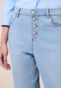 Los jeans de mezclilla azul claro presentan un diseño de cintura alta con cinco botones, incluyendo acentos florales decorativos. Textura suave y forma ajustada.