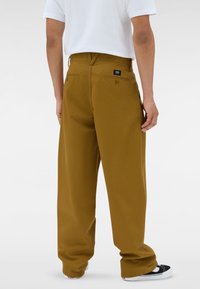 Persona vestita con pantaloni a gamba larga color senape, t-shirt bianca e sneaker bianche e nere, rivolto verso il muro bianco uniforme.