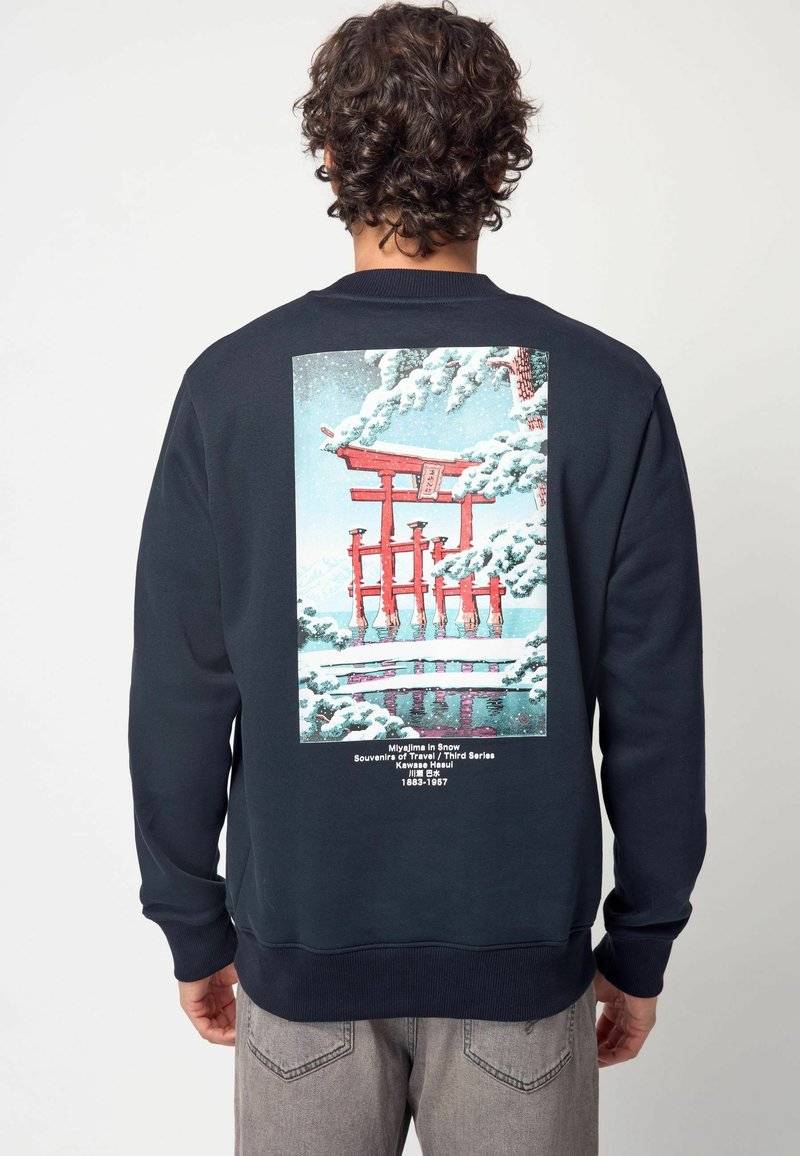 Felpa blu marino con un'illustrazione grafica sulla schiena, che presenta un cancello torii rosso in un paesaggio innevato, con del testo sotto l'opera.