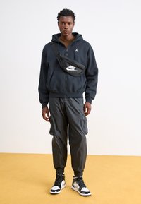 Sudadera con capucha negra con cremallera y logo, pantalones cargo oscuros con puños elásticos y zapatillas negras y blancas. Los accesorios incluyen una riñonera negra.