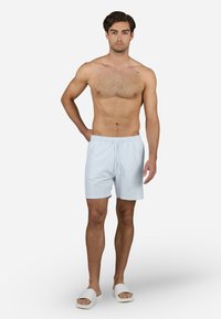 Lyseblå badebukser med elastisk liv og snor, laget av glatt stoff. Brukt med hvite sandaler med åpen tå.
