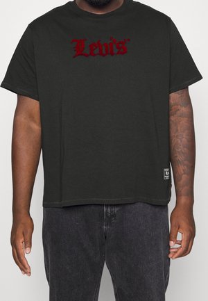 Czarna koszulka Levi's z czerwonym logo w stylu gotyckim na piersi, noszona przez osobę z tatuażem na prawym przedramieniu i ciemnymi dżinsami.