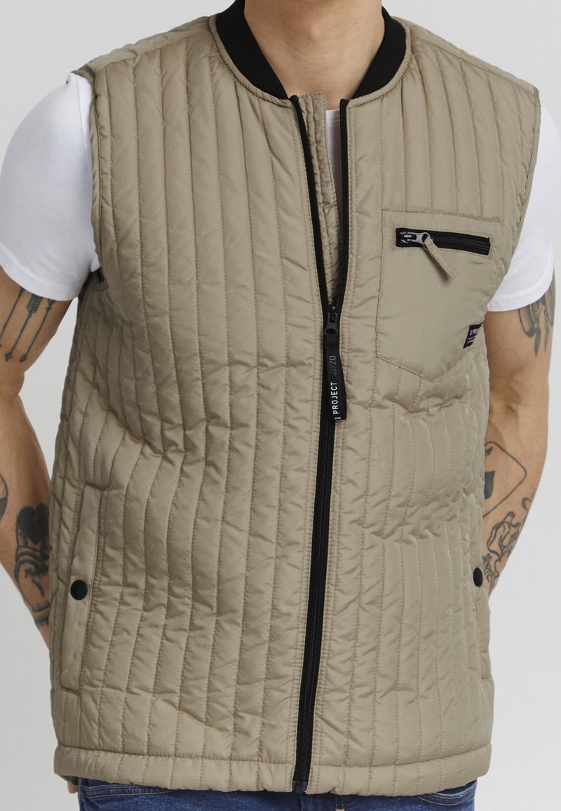 Gilet imbottito beige con zip frontale nera, colletto corto e tasca sul petto; presenta cuciture orizzontali e dettagli con bottoni a scatto.