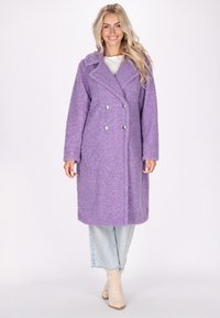 Cappotto doppio petto viola, strutturato, con grandi bottoni argentati, colletto a ponte e maniche lunghe, abbinato a jeans chiari e stivaletti beige alla caviglia.