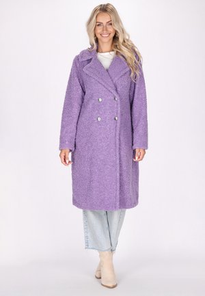 Cappotto doppio petto viola, strutturato, con grandi bottoni argentati, colletto a ponte e maniche lunghe, abbinato a jeans chiari e stivaletti beige alla caviglia.
