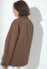 Woman with short dark hair wearing a loose brown coat, standing sideways against a white background casting a shadow.