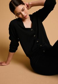Cardigan negru cu nasturi la față, decolteu în formă de v, și manșete cu nervuri, purtat peste pantaloni negri asortați, pe un fundal bej deschis.