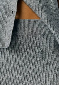 Graues geripptes Strickkleidungsstück mit Knopfverschluss und Ausschnittdetail, das einen strukturierten Stoff mit vertikalen Streifen zeigt.