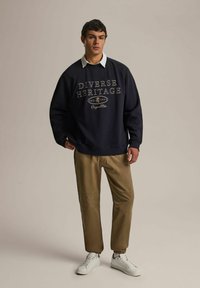 Navy bluza z napisem "Różnorodne Dziedzictwo", beżowe spodnie i białe sneakersy. Posiada ribbę na mankietach i u dołu, luźny krój oraz casualowy design.
