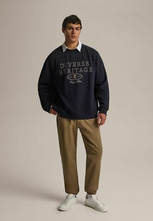 Navy bluza z napisem "Różnorodne Dziedzictwo", beżowe spodnie i białe sneakersy. Posiada ribbę na mankietach i u dołu, luźny krój oraz casualowy design.