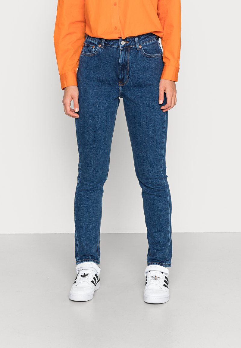 Calças skinny de denim azul com uma cintura alta, cinco bolsos e costura contrastante, combinadas com ténis brancos e uma camisa laranja.