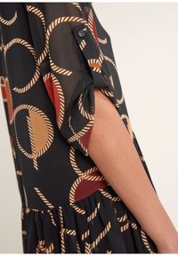 Arm visad i en svart genomskinlig klänning med veckad ärm och geometriskt cirkelmönster i brunt, beige och vitt repdesign.