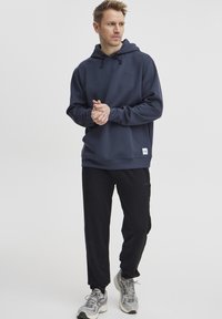 Jūras spēku kapuce ar priekšējo kabatu, apvienota ar melniem biksēm un pelēkiem sporta apaviem. Audums izskatās mīksts un tam ir brīvs piegriezums.