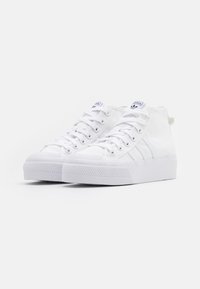 Zapatillas altas blancas con parte superior de lona, suela de goma, punta texturizada y tres rayas blancas en el lado. Disponen de cordones planos y una pestaña en el talón.