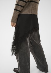 Jupe asymétrique noire avec bordure en dentelle, associée à un jean gris à jambes larges. Le pull est beige avec des rayures noires, présentant une texture côtelée.