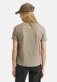 Khaki bomulds t-shirt med korte ærmer og rund hals, parret med en brun fløjl baseball cap, set fra ryggen.