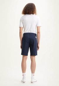 Osoba s kudrnatými vlasy, oblečená v bílém tričku, námořnických modrých šortkách, bílých sockách a bílých teniskách, stojí otočená zády.