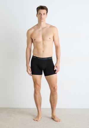 Man standing barefoot against white wall wearing black Tommy Hilfiger boxer briefs, facing forward with neutral expression.