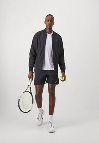 Must Nike bomberjakk valge polo, mereväe lühikeste pükste ja valgete tossude peal. Hoidmas tennise reketit ja rohelist tennispalli.