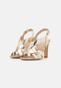 Sandales à talons hauts en métal doré avec des brides croisées, bouts ouverts et boucles réglables autour de la cheville sur un fond blanc.