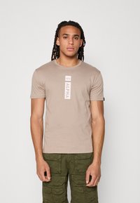 Beige T-shirt med rund halsudskæring og korte ærmer, med et vertikalt "ALPHA" logo. Parret med olivengrønne cargo shorts med flere lommer.