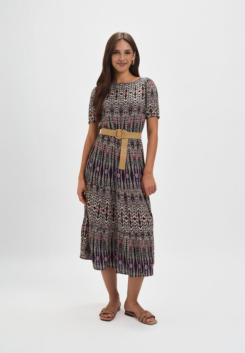 Maxi sukienka z czarno-biało-czerwonym wzorem, krótkimi rękawami, dopasowaną talią z beżowym paskiem oraz płaskimi sandałami.