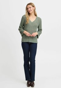 Grön stickad tröja med V-hals, utsvängda ärmar och öppna vävmönster, kombinerad med mörkblå jeans och bruna boots med spetsig tå.