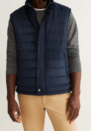 Homme portant un gilet matelassé sans manches bleu marine sur une chemise à manches longues grise et un pantalon beige, debout devant un fond uni.