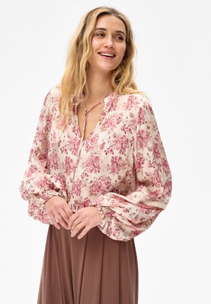 Vrouw met lang blond haar, gekleed in een crème blouse met roze bloemendessin en ballonmouwen, gecombineerd met een bruine rok, glimlachend en naar rechts kijkend.
