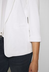 Bílý pruhovaný blazer s jednou přední kapsou a ohrnutými rukávy, zkombinovaný s tmavými džíny. Hladká textilie a přiléhavý střih.