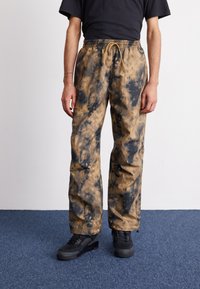 Tan and black tie-dye pants with an elastic waistband and drawstring. Made of a lightweight fabric, featuring side pockets and a relaxed fit.