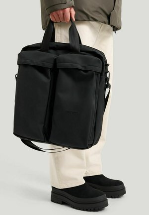 Person holding black Tretorn bag with two front pockets, wearing beige pants and black platform shoes against a plain background.