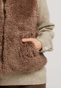 Écharpe en fausse fourrure marron avec une apparence texturée et duveteuse, portée sur un pull gris clair avec des poignets boutonnés.