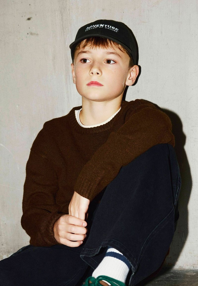 Jeune garçon portant une casquette noire et un pull marron, assis contre un mur clair, regardant sur le côté avec une expression pensive.